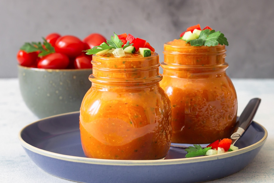 Gazpacho recept Stämningsbild: Två skruvförsedda burkar fyllda med röd gazpacho arrangerade på en blå tallrik, i bakgrunden en skål med tomater